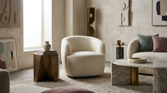 River cream white boucle barrel swivel chair in a light contemporary living room, angled between a window and a sofa with a marble coffee table alongside.
