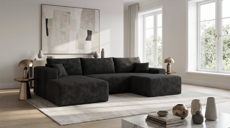 A black corduroy cloud sectional sofa shown at true scale in a bright, airy contemporary living room with large windows and a light area rug