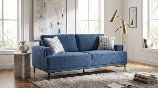Blue chenille sofa with loose cushions and patterned pillows in a sunlit living room with tall windows