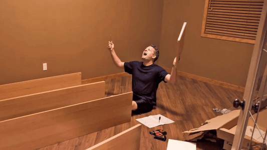 Frustrated man sitting on the floor surrounded by furniture pieces, expressing exasperation during assembly.