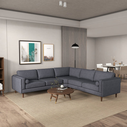 A modern living room showcases the Ashcroft Furniture Amber MCM Corner Sectional Sofa 103 in gray on a beige rug, paired with a round wooden coffee table, wall art, floor lamp, and a dining area with a white table and chairs in the background.