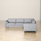 The Amber MCM Corner Sectional Sofa 103 by Ashcroft Furniture, shown in modern light gray with wooden legs, is placed on a light wood herringbone floor against a plain beige wall.