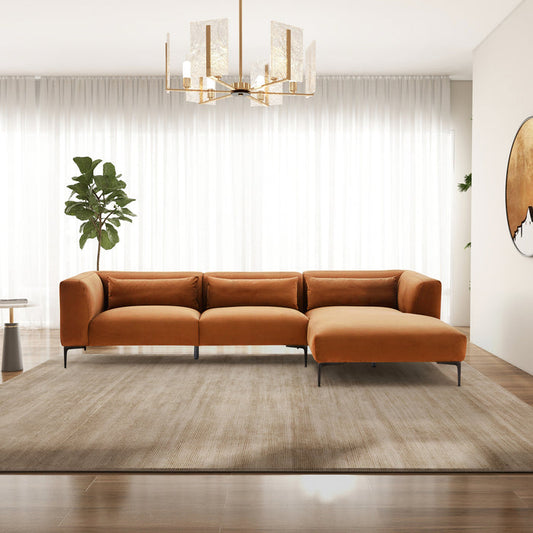 A modern living room featuring the Ashcroft Furniture Laley MCM Right-Facing Chaise Sofa Sectional 122 in orange, set on a beige rug with wooden floors, a large window with sheer curtains, potted plant, wall art, and a gold chandelier.