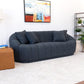 A modern living room showcases the Ashcroft Furniture Maximilian Japandi Channel Tufted Boucle Sofa 86” in dark blue with two matching pillows, a tan ottoman, white textured rug, abstract painting, and black metal shelf with plants.