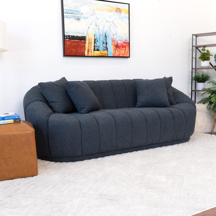 A modern living room showcases the Ashcroft Furniture Maximilian Japandi Channel Tufted Boucle Sofa 86” in dark blue with two matching pillows, a tan ottoman, white textured rug, abstract painting, and black metal shelf with plants.