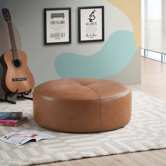 The Ashcroft Furniture Co. Rochelin Round Tan Leather Ottoman 36 with genuine leather upholstery sits on a white patterned rug, near an acoustic guitar, a book, and framed wall art in a sunlit mid-century modern room.