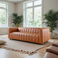 Elrosa Channel Tufted Cognac Faux Leather Sofa in a bright living room with large windows and indoor plants.