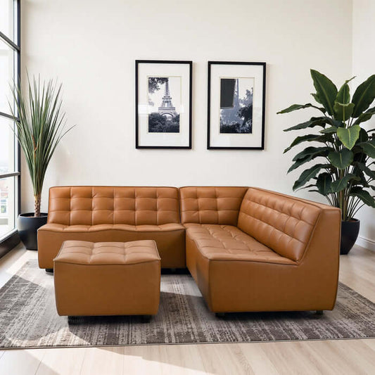 A modern living room featuring the Ashcroft Furniture Co Selen MCM Leather Modular Corner Sofa Sectional with Ottoman (91W) in tan vegan leather on a patterned rug, two framed black-and-white photos, and tall green plants by large windows.