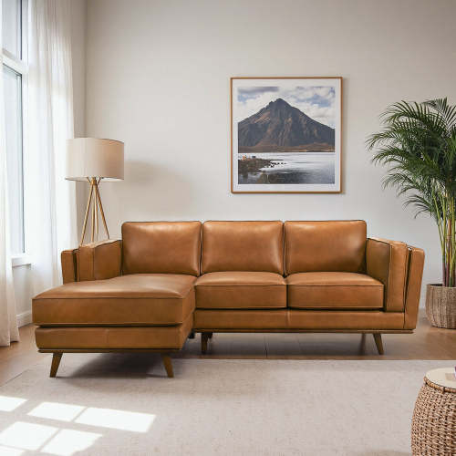 A modern living room features the Chase MCM Tan Leather Chaise Sofa Sectional 92 by Ashcroft Furniture, a floor lamp, a large potted plant, and a framed mountain landscape photo. Sunlight streams through sheer white curtains at the window.