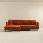 The Ashcroft Furniture Blake Velvet Upholstered L-Shape Chaise Sofa Sectional 107 in plush orange velvet, featuring a reversible chaise and four matching cushions, sits against a light beige wall in a minimalist room.