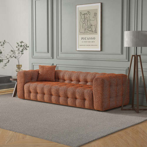 A modern living room with a glam vibe showcases the Ashcroft Furniture Eden Modern Boucle Bubble Sofa 90 in orange, complemented by a gray area rug, tall floor lamp, small plant on a side table, and Picasso art on the paneled gray wall.