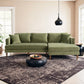 A modern living room featuring the Ashcroft Furniture Blake Velvet Upholstered L-Shape Chaise Sofa Sectional 107 in green, two throw pillows, a black round coffee table with a wooden fruit tray, patterned rug, black pendant light, and large windows for natural light.