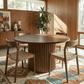 Merlin 35" Solid Wood Round Dining Table in Walnut with Ribbed Pedestal Base, surrounded by four woven chairs in a cozy dining room.