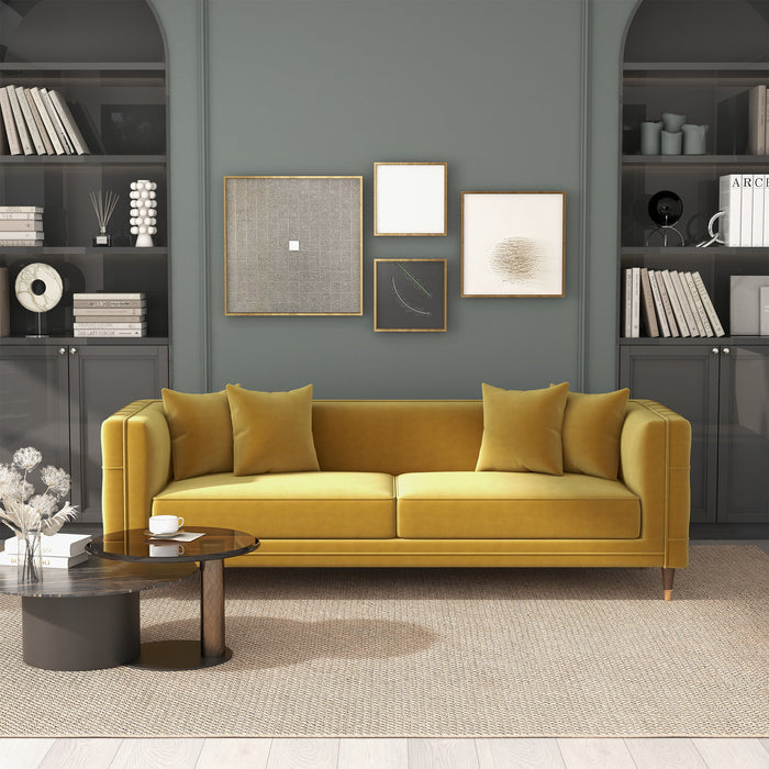 A modern living room featuring the Ashcroft Furniture Edward MCM Elegant Tufted Velvet Sofa 90 in mustard yellow velvet, four matching pillows, a round coffee table, built-in dark shelves with books and decor, and framed art on a greenish-gray wall.