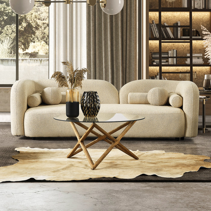 A modern living room featuring the Donna Minimalist Curved Boucle Sofa 93 by Ashcroft Furniture in cream, paired with a glass coffee table with wooden base, decorative vases, beige rug, bookshelf with ambient lighting, and a large sunlit window.
