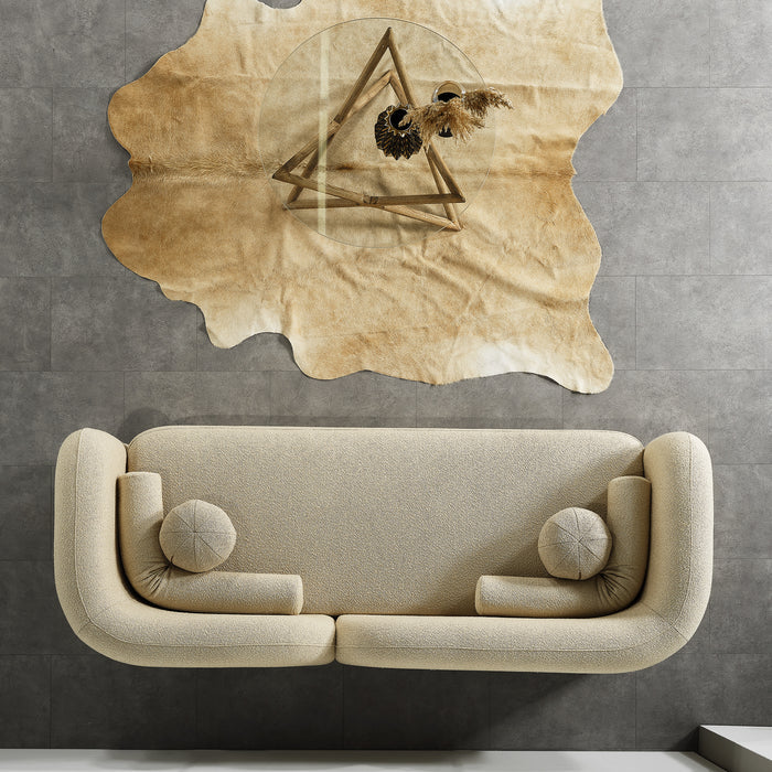 The Donna Minimalist Curved Boucle Sofa 93 by Ashcroft Furniture in beige, accented with round cushions, sits against a gray tiled wall topped by a tan animal hide with geometric triangle and feather artwork.