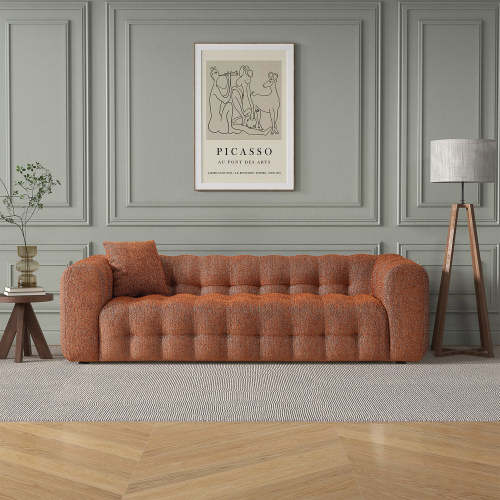 A modern living room with the Ashcroft Furniture Eden Modern Boucle Bubble Sofa 90 in tufted orange, accented by a side table with plant, floor lamp, and Picasso print on gray paneled walls. Wood floors and a large rug complete the glam look.