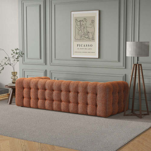 A modern living room showcasing the 90 Eden Modern Boucle Bubble Sofa by Ashcroft Furniture in orange, set against a gray paneled wall with framed Picasso art, a gray-shaded floor lamp, a side table with branches, and a textured gray rug.