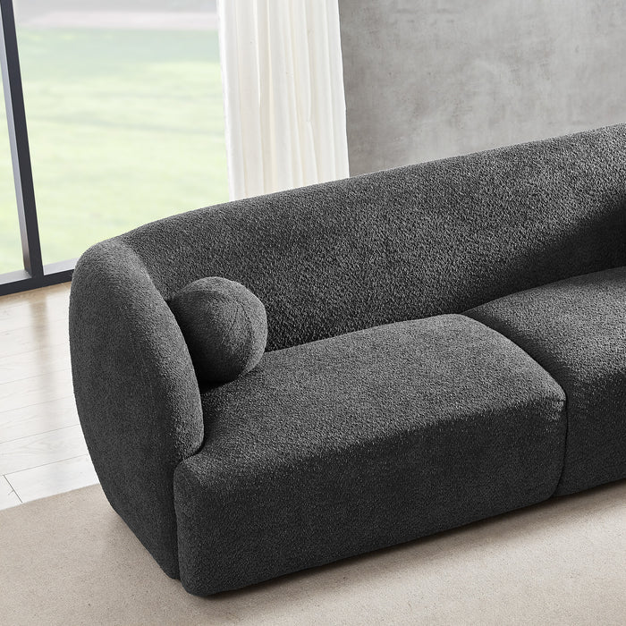 The Anna Boucle Fabric Upholstered Sofa 87 by Ashcroft Furniture, shown in dark gray with textured boucle fabric and a matching round cushion, sits in a bright room with light wood floors, a beige rug, and floor-to-ceiling windows.