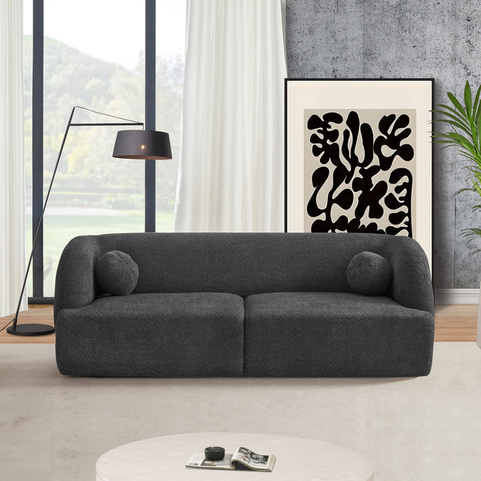 A modern living room featuring the Ashcroft Furniture Anna Boucle Fabric Upholstered Sofa 87 in dark gray with two round cushions, a black floor lamp, abstract black-and-white art, a green plant, and large windows with white curtains.