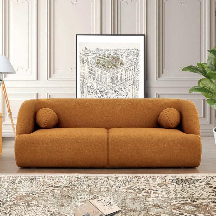 A modern living room with the Anna Boucle Fabric Upholstered Sofa 87 by Ashcroft Furniture in brown, two round cushions, cityscape artwork on a paneled wall, a white floor lamp, green plant, and a patterned rug.