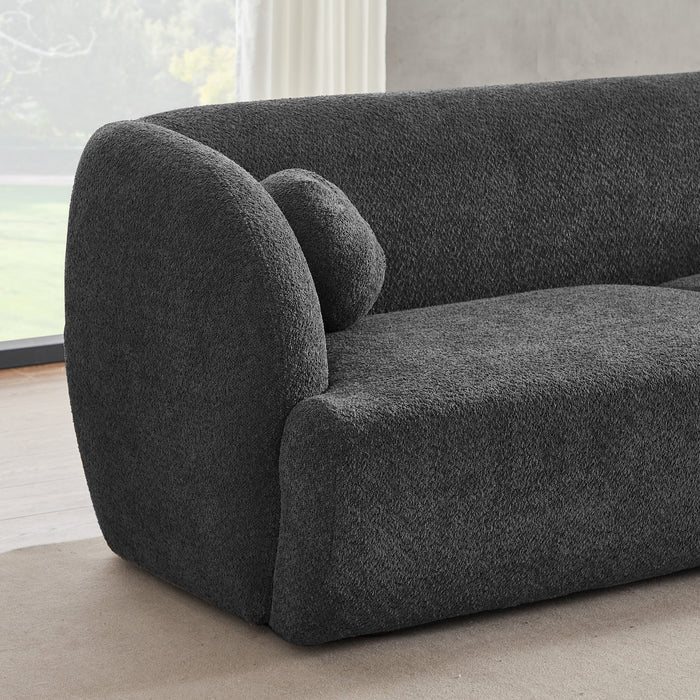 The Anna Boucle Fabric Upholstered Sofa 87 by Ashcroft Furniture, shown in modern dark gray with textured boucle fabric, rounded armrests, and a matching cushion, is placed on a light rug near a large window with sheer white curtains.