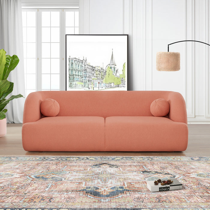 A modern living room showcases the Anna Boucle Fabric Upholstered Sofa 87 in peach by Ashcroft Furniture, paired with two round pillows, a patterned rug, potted plant, floor lamp, and cityscape art against white walls with tall windows.