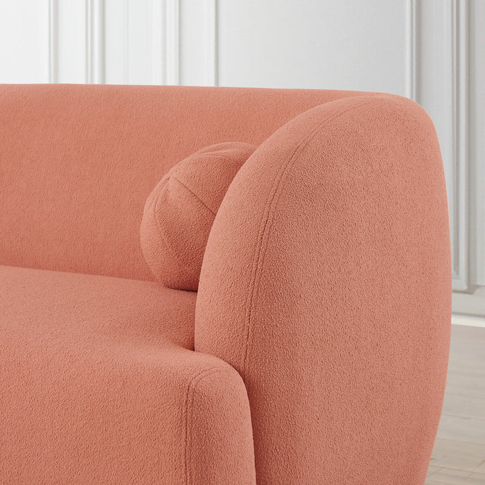 Close-up of the Anna Boucle Fabric Upholstered Sofa 87 in coral from Ashcroft Furniture, featuring a modern curved design with a matching round pillow—an elegant addition to any light-paneled room with wooden floors.