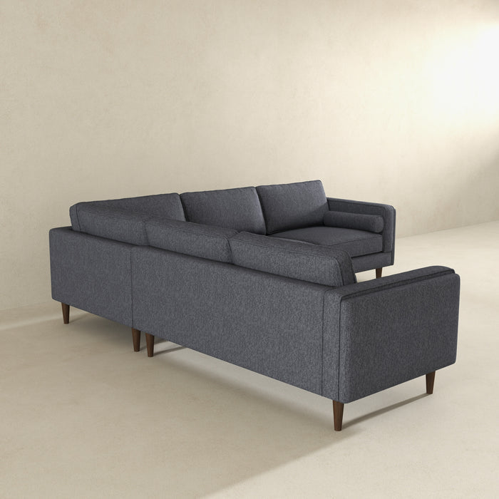 The Amber MCM Corner Sectional Sofa 103 in dark gray by Ashcroft Furniture, featuring wooden legs, is set in a bright minimalist room with light beige walls and floor, shown from the back and side.