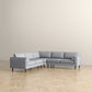 The Ashcroft Furniture Amber MCM Corner Sectional Sofa 103 in light gray, featuring wooden legs, is arranged on a light wood herringbone floor beside a plain beige wall.