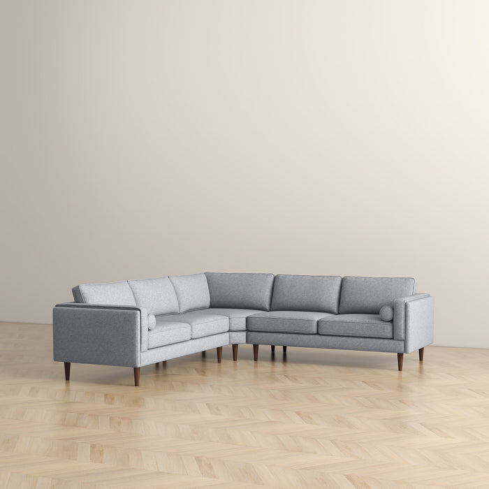 The Ashcroft Furniture Amber MCM Corner Sectional Sofa 103 in light gray, featuring wooden legs, is arranged on a light wood herringbone floor beside a plain beige wall.