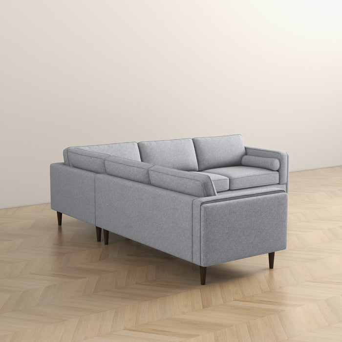 The Amber MCM Corner Sectional Sofa 103 by Ashcroft Furniture in light gray features clean lines and tapered wooden legs. Shown on a herringbone floor against an off-white wall, it highlights the back and side’s premium craftsmanship.