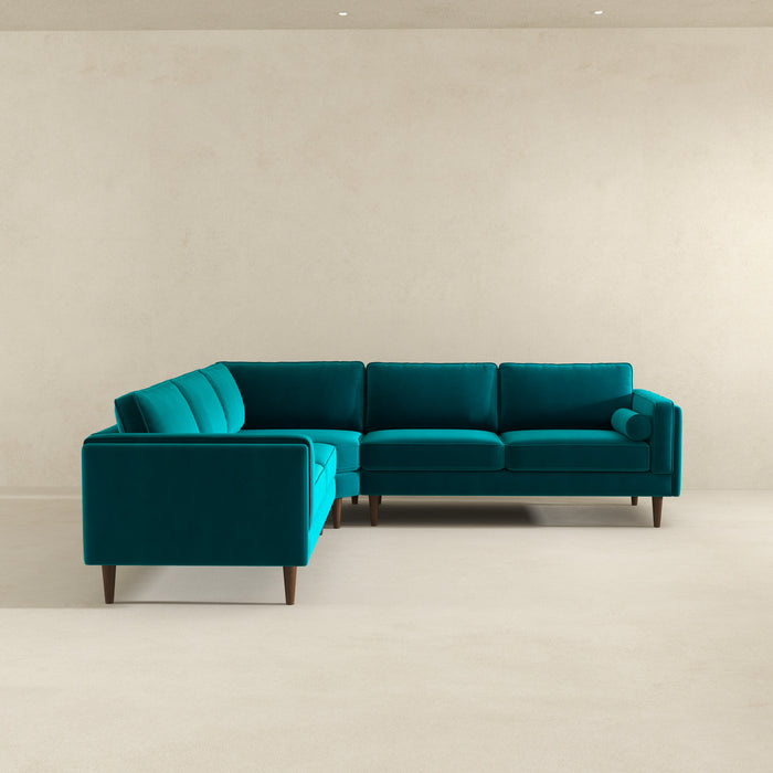 The Ashcroft Furniture Amber MCM Corner Sectional Sofa 103 in teal stands out in an empty room with beige walls and floor. A cylindrical cushion on one end emphasizes its premium craftsmanship and stylish wooden legs.