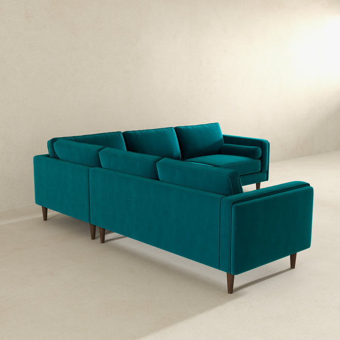 The Amber MCM Corner Sectional Sofa 103 by Ashcroft Furniture, shown in modern teal with plush cushions and wooden legs, sits stylishly in a minimalist room featuring light beige walls and floors. Premium craftsmanship is visible from the angled back view.