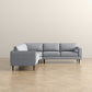 The Ashcroft Furniture Amber MCM Corner Sectional Sofa 103 in light gray features three seat and back cushions plus wooden legs, shown on a light wood herringbone floor against a plain beige wall.
