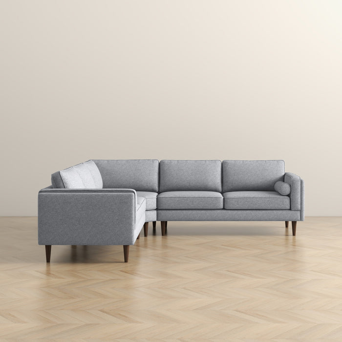 The Ashcroft Furniture Amber MCM Corner Sectional Sofa 103 in light gray features three seat and back cushions plus wooden legs, shown on a light wood herringbone floor against a plain beige wall.