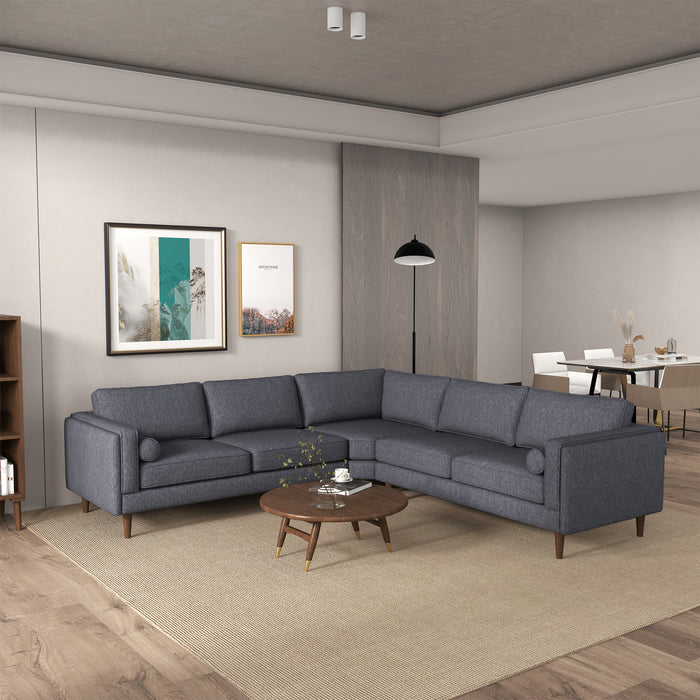 A modern living room showcases the Ashcroft Furniture Amber MCM Corner Sectional Sofa 103 in gray on a beige rug, paired with a round wooden coffee table, wall art, floor lamp, and a dining area with a white table and chairs in the background.