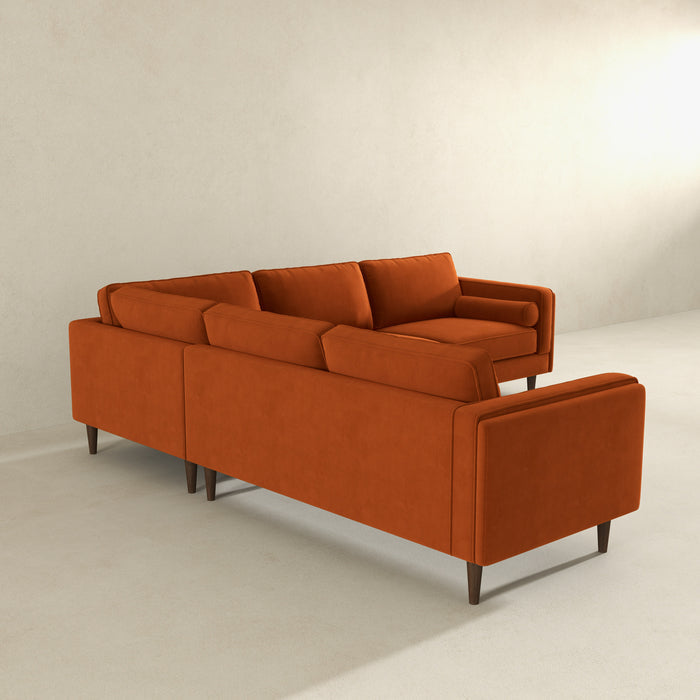 The Amber MCM Corner Sectional Sofa 103 by Ashcroft Furniture, shown in burnt orange with clean lines and wooden legs, brings modern style and premium craftsmanship to a minimalist room with light walls and flooring.