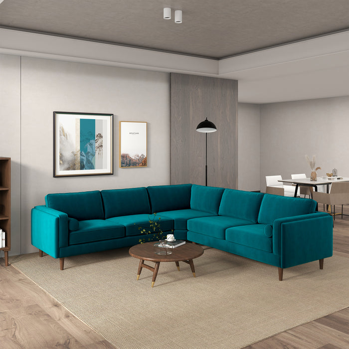 A modern living room features the Ashcroft Furniture Amber MCM Corner Sectional Sofa 103 in teal, arranged in an L-shape with a round wooden coffee table and neutral walls. Two framed artworks hang above, a floor lamp is in the corner, and a dining area is visible beyond.