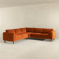 The Amber MCM Corner Sectional Sofa 103 by Ashcroft Furniture, shown in burnt orange, features clean lines and wooden legs, adding a modern touch to a minimalist light beige room with smooth floors and plain walls.