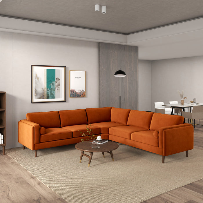 A modern living room featuring the Ashcroft Furniture Amber MCM Corner Sectional Sofa 103 in orange on a beige rug, paired with a round wooden coffee table, wall art, wood floors, neutral walls, and an open dining area in the background.