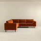 The Amber MCM Corner Sectional Sofa 103 by Ashcroft Furniture, shown in vibrant orange velvet with wooden legs, highlights premium craftsmanship against a plain off-white wall and light floor in a minimal, empty room.