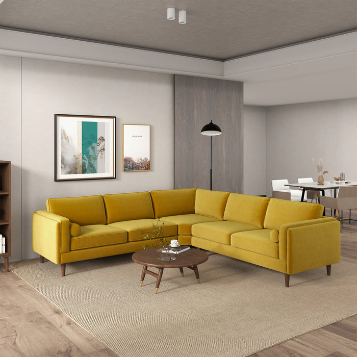 A modern living room featuring the Amber MCM Corner Sectional Sofa 103 in mustard yellow by Ashcroft Furniture, arranged L-shaped with a round wood coffee table, beige rug, wall art, floor lamp, and dining area visible in the background.