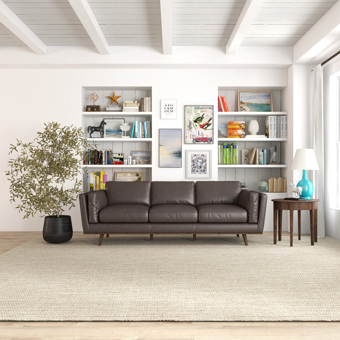 A modern living room features the Ashcroft Furniture Chase MCM Genuine Leather Sofa 91 in dark brown, a large potted plant, side table with lamp, and built-in shelves. Light walls and a woven rug enhance the airy feel.