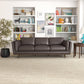 A modern living room showcases the Ashcroft Furniture Chase MCM Genuine Leather Sofa 91 in dark brown, placed before white built-in bookshelves with colorful books, art, and decor. A round wooden side table with a lamp and large plant completes the mid-century look.