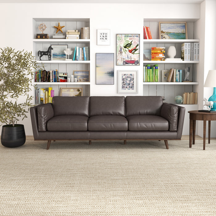 A modern living room showcases the Ashcroft Furniture Chase MCM Genuine Leather Sofa 91 in dark brown, placed before white built-in bookshelves with colorful books, art, and decor. A round wooden side table with a lamp and large plant completes the mid-century look.