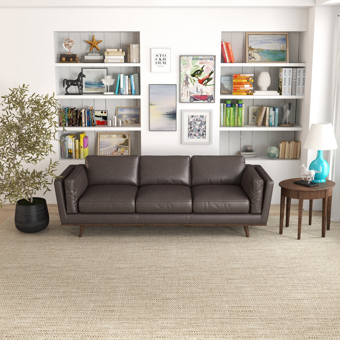A modern living room featuring the Ashcroft Furniture Chase MCM Genuine Leather Sofa 91, a beige woven rug, round wooden side table with turquoise lamp, potted plant, and white bookshelves filled with books and décor for mid-century flair.