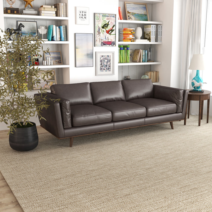 A living room features the Ashcroft Furniture Chase MCM Genuine Leather Sofa 91 on a beige rug, with a round wood side table, white lamp, potted plant, and shelves displaying books, art, and decor.
