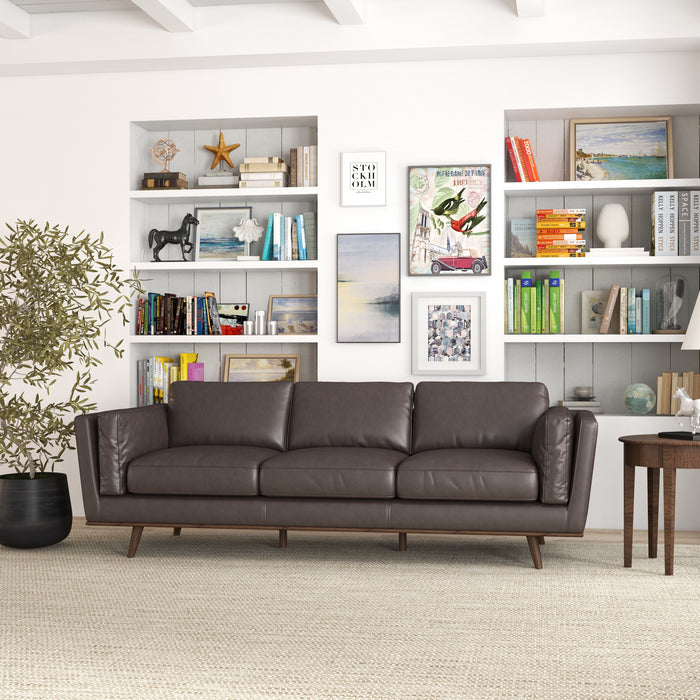 A modern living room featuring the Ashcroft Furniture Chase MCM Genuine Leather Sofa 91 in dark brown, a large leafy plant, a side table with a lamp, and built-in shelves with books and decor against a white wall.