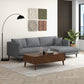 A modern living room featuring the Ashcroft Furniture Blake Velvet Upholstered L-Shape Chaise Sofa Sectional 107, paired with a wooden coffee table, black arc floor lamp, wall panels, potted plant, large window, light rug, and wood flooring.